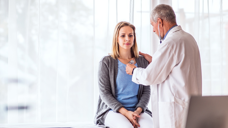 Senior white male doctor examining white woman with blonde hair, listening to chest with stethoscope